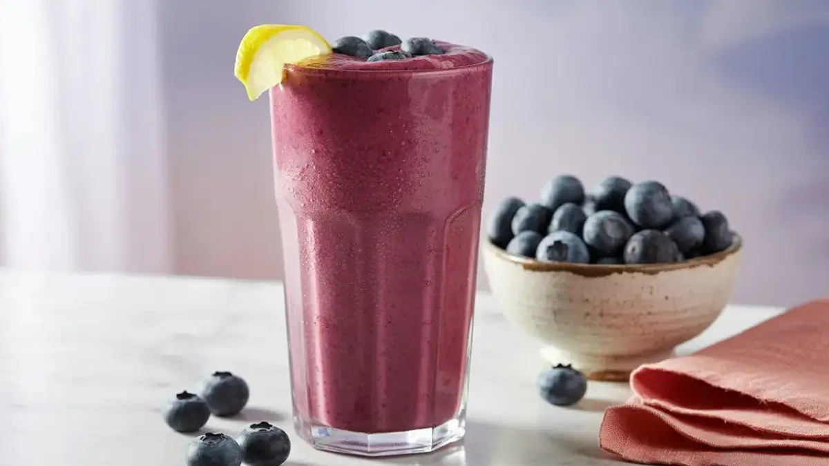 Tall glass of vibrant purple lemon-blueberry smoothie, garnished with fresh blueberries and a lemon wedge, with a bowl of blueberries and a coral napkin in the background.