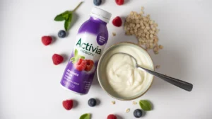 Flat lay of Activia probiotic drink bottle next to a bowl of yogurt, berries, oats, and mint on white background
