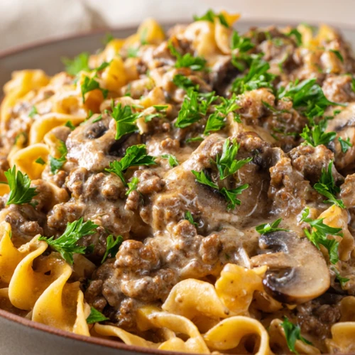 Creamy ground beef stroganoff served in a shallow bowl with mushrooms, parsley, and egg noodles on a neutral linen background