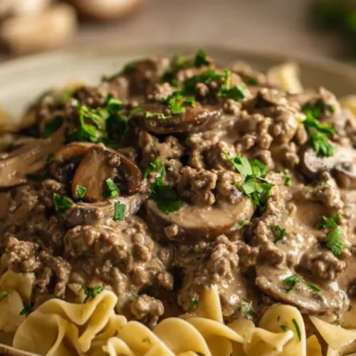 A close-up of creamy ground beef stroganoff with sautéed mushrooms and fresh parsley served over egg noodles on a rustic ceramic plate