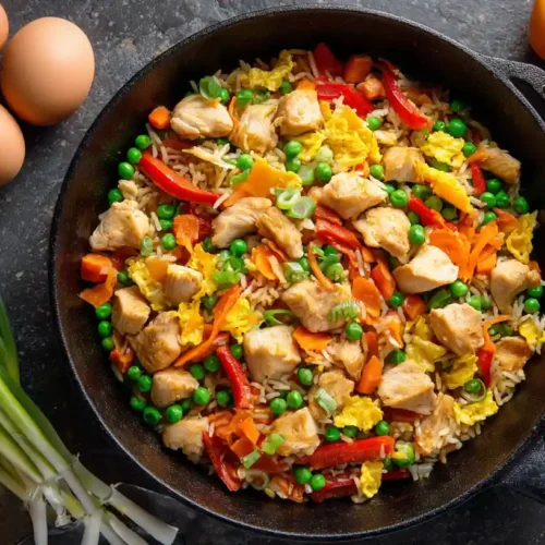 Overhead view of homemade healthy chicken fried rice with lean chicken, peas, carrots, and bell peppers in a cast-iron wok, surrounded by fresh eggs and scallions