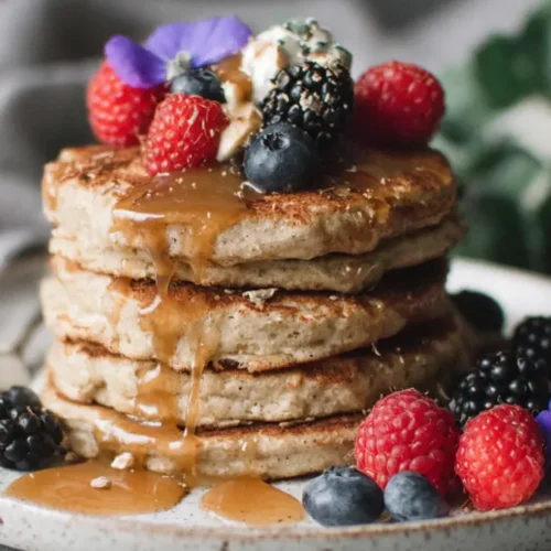 Stack of banana cottage cheese pancakes topped with raspberries blueberries and blackberries, maple syrup dripping, on a speckled ceramic plate in soft light