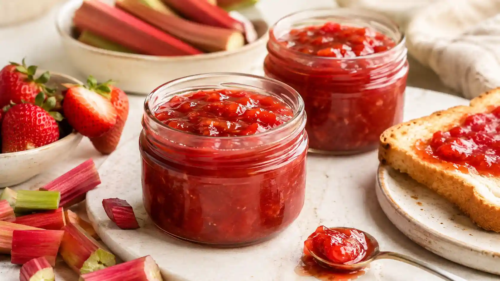 Strawberry rhubarb jam in glass jars with strawberries and rhubarb stalks on a light table.