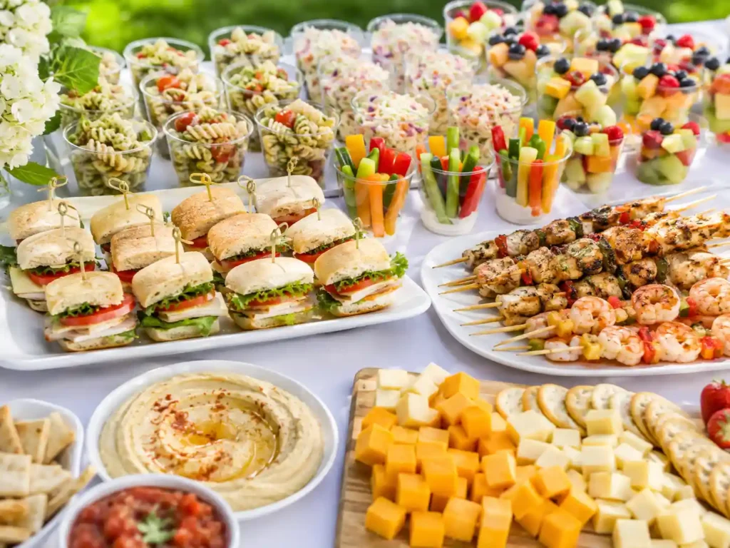 Summer buffet table with mini sandwiches, skewers, dips, pasta cups, slaw cups, and fruit cups