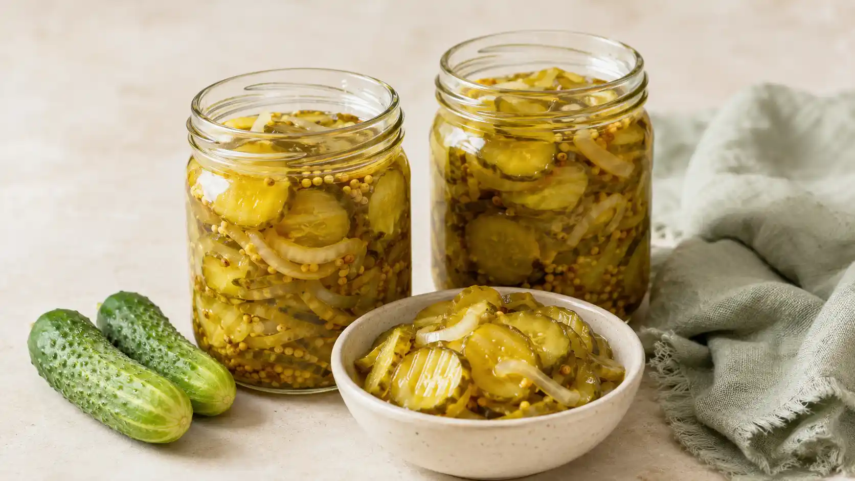 Two jars and a cream bowl filled with homemade bread and butter pickle chips, sliced onion, and mustard seeds.