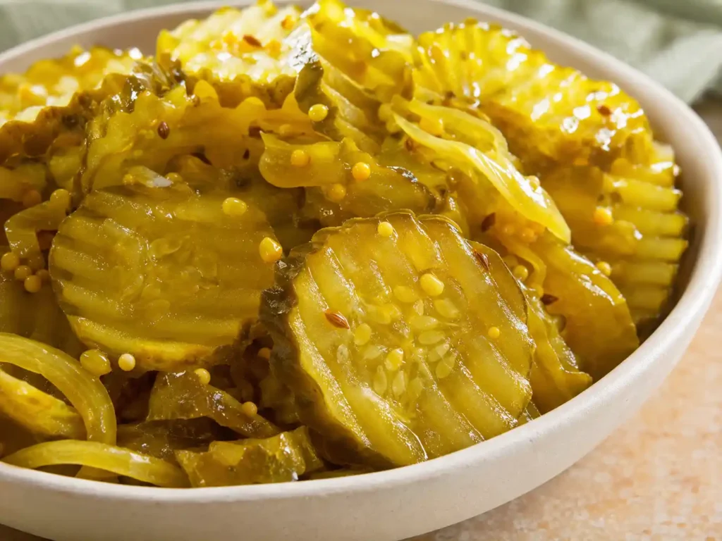 Sharp close-up of golden bread and butter pickle chips with onion, mustard seeds, celery seeds, and glossy brine.