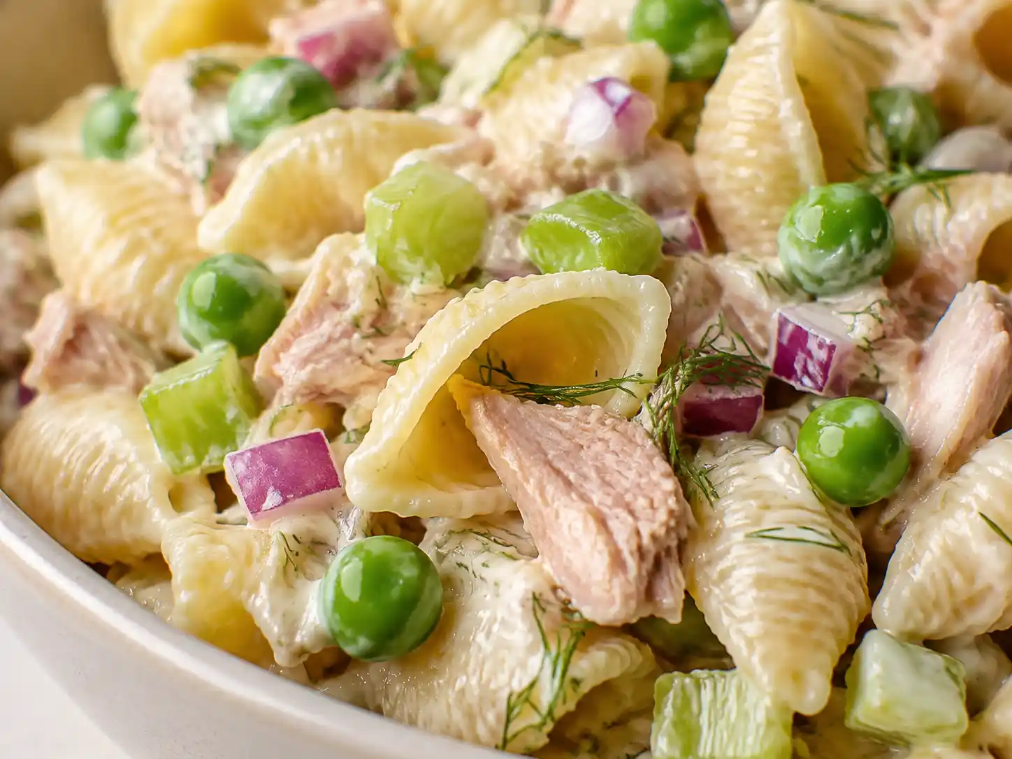 Close-up of creamy dressing clinging to shell pasta with tuna, peas, celery, red onion, and dill